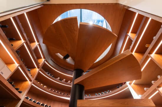 Interior view of a luxury spiral wine cellar with curved cherry wood shelving and a sculptural central staircase, illuminated by integrated LED lighting and natural light from a circular glass ceiling.